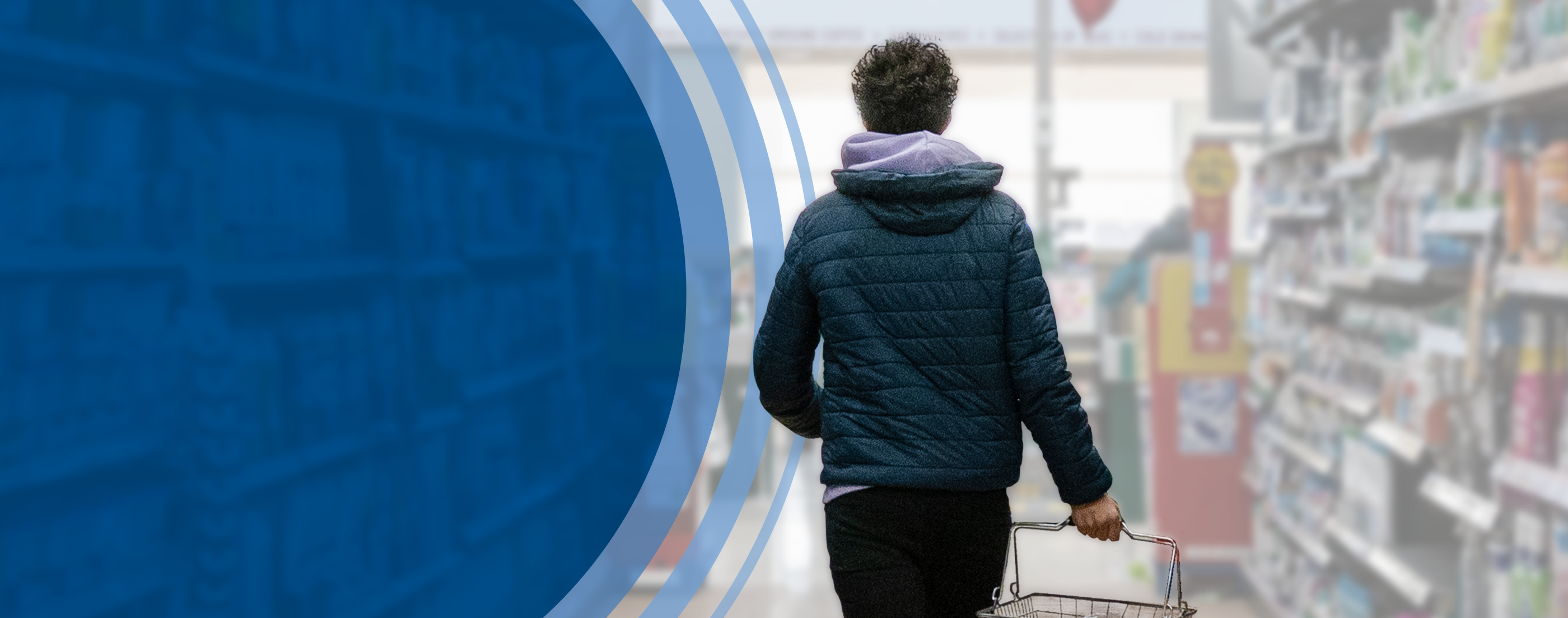 A person in a dark blue, puffy hooded jacket walks away from the camera and down a supermarket aisle with household items stacked on the shelves on both sides. The person carries a shopping basket containing only the few items they could afford as they walk toward the checkout lanes.