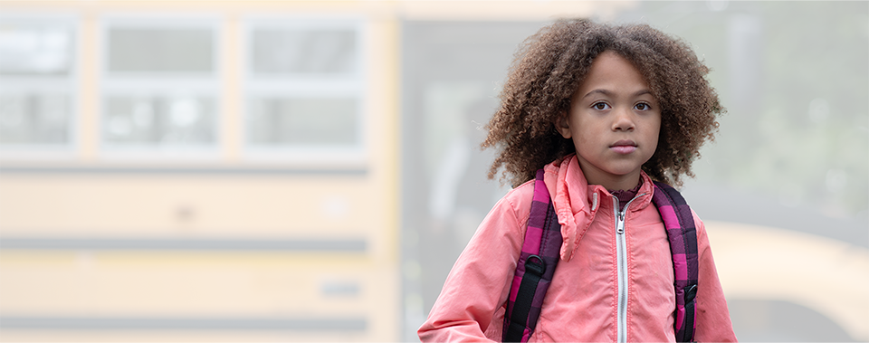 A school-age child, wearing a backpack and a pink jacket, stands in front of a school bus and stares at the camera with a serious expression.