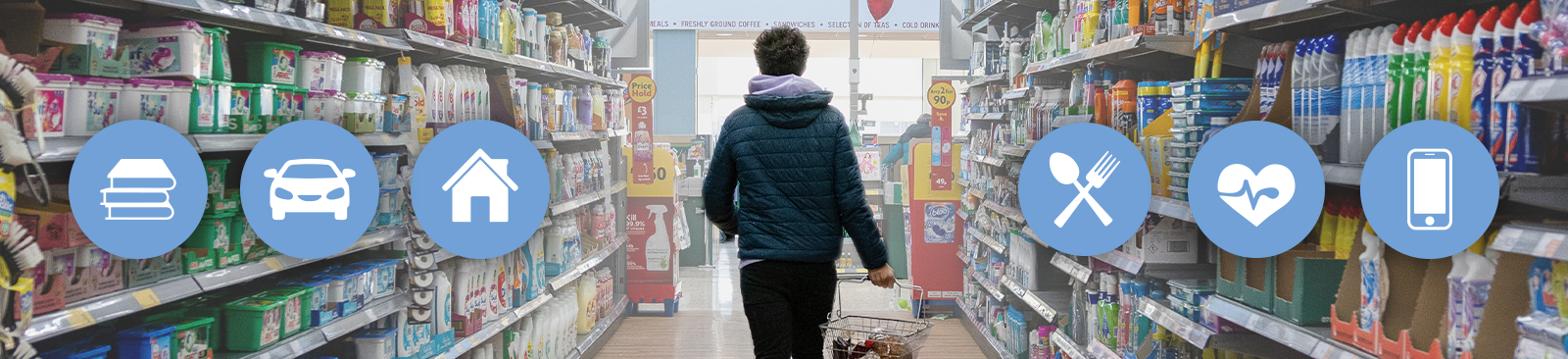 A person in a dark blue, puffy hooded jacket walks away from the camera and down a supermarket aisle with household items stacked on the shelves on both sides. The person carries a shopping basket containing only the few items they could afford as they walk toward the checkout lanes.