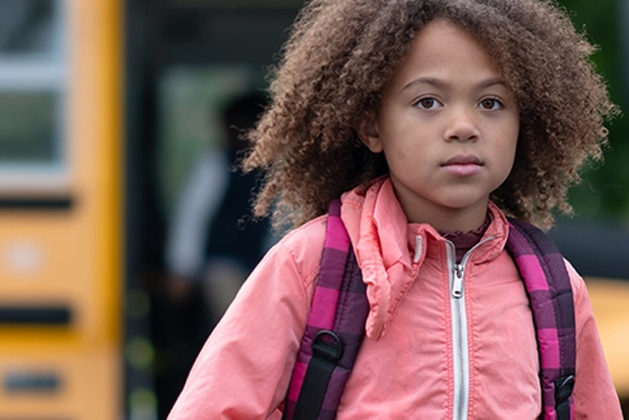 A school-age child, wearing a backpack and a pink jacket, stands in front of a school bus and stares at the camera with a serious expression.